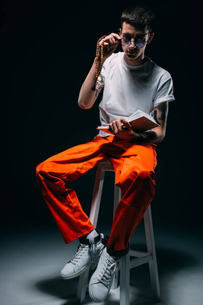 Man in orange uniform with cuffs on legs reading bible while sitting on stool on dark background