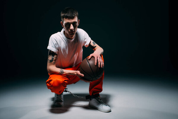 Man in orange pants and cuffs holding basketball ball on dark background