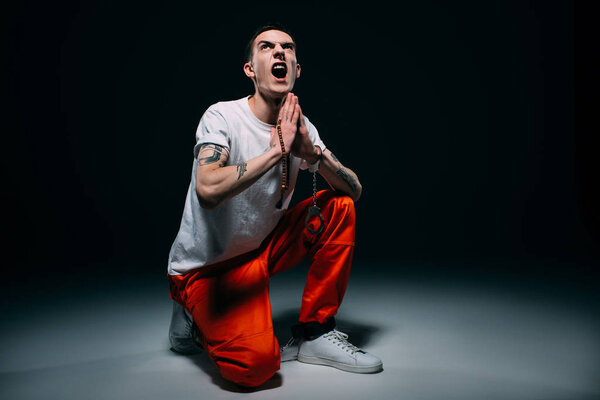 Screaming man in prison uniform and cuffs holding rosary and praying on dark background