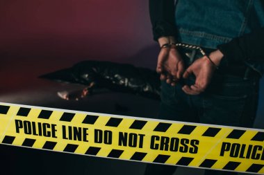 Rear view of man in cuffs by dead body behind yellow tape on dark background clipart