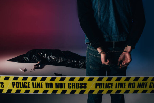 Man in cuffs by dead body behind yellow line on dark background