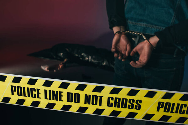 Rear view of man in cuffs by dead body behind yellow tape on dark background