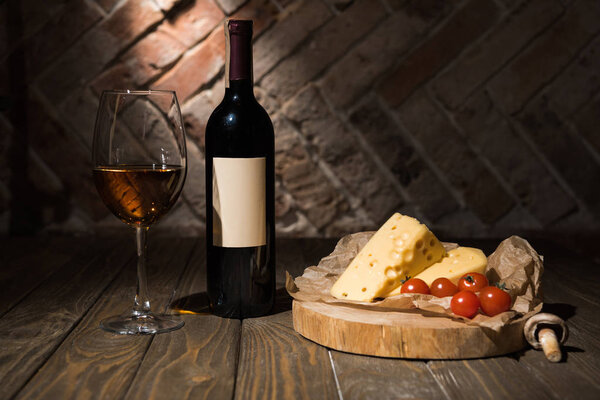 close up view of bottle and glass of wine with cheese and cheery tomatoes on baking paper on wooden decorative stump