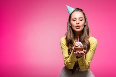attractive woman with party hat holding cupcake and blowing out candle isolated on pink clipart