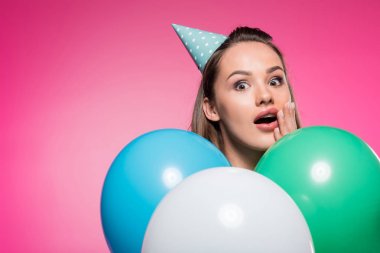 shocked attractive woman with party hat and balloons isolated on pink clipart