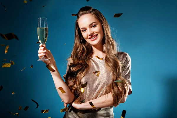 笑容迷人的女人站在玻璃香槟下落在蓝色的五彩纸屑 — 图库照片