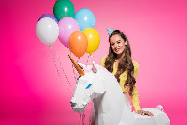attractive woman with party hat, balloons and unicorn toy isolated on pink