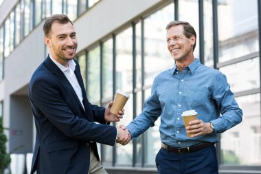 smiling businessmen with disposable cups of coffee shaking hands near office building clipart