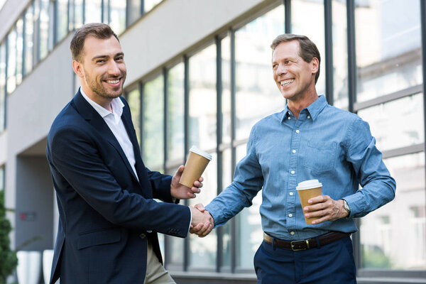smiling businessmen with disposable cups of coffee shaking hands near office building