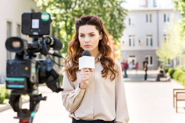 serious anchorwoman with microphone standing in front of digital video camera