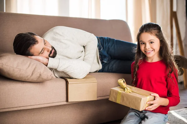 Fille tenant boîte cadeau tandis que père dormant sur canapé — Photo de stock