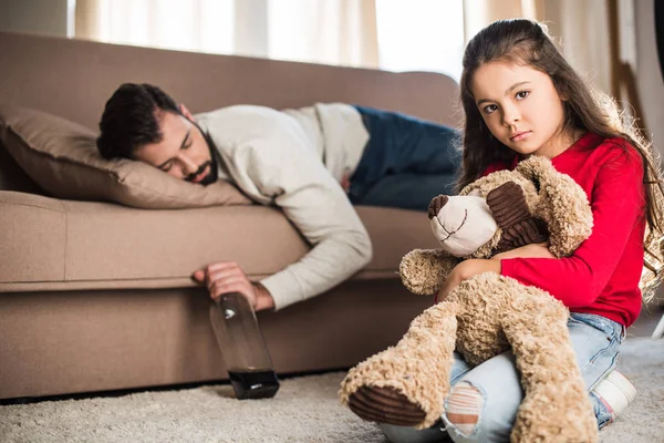 Грустная дочь сидит с плюшевым мишкой пока пьяный отец спит на диване — стоковое фото