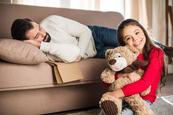 Padre durmiendo en sofá e hija sentado con osito de peluche - foto de stock