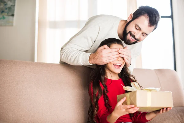Padre sonriente cubriendo los ojos de la hija desde atrás y presentando regalo - foto de stock