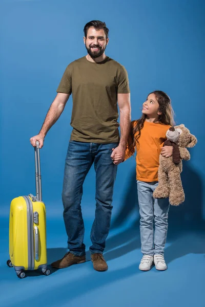 Padre e hija de pie con bolsa de viaje amarilla y oso de peluche en azul - foto de stock