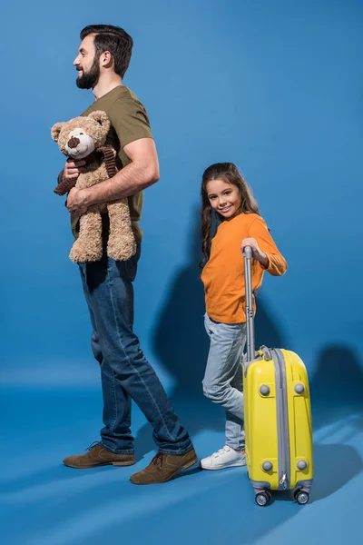 Padre con osito de peluche e hija con bolso sobre ruedas en azul - foto de stock