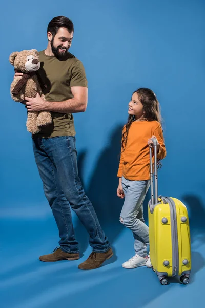 Father with teddy bear and daughter with bag on wheels looking at each other on blue — Stock Photo