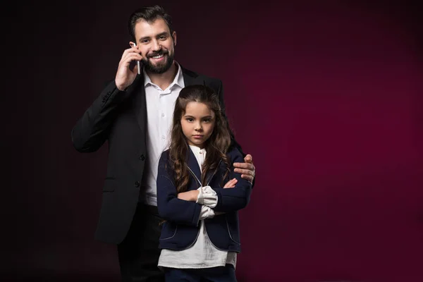 Padre hablando por teléfono inteligente y abrazando hija seria con las manos cruzadas en color burdeos - foto de stock