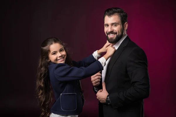 Улыбающаяся дочь чинит галстук отца и смотрит в камеру на бордовом — стоковое фото