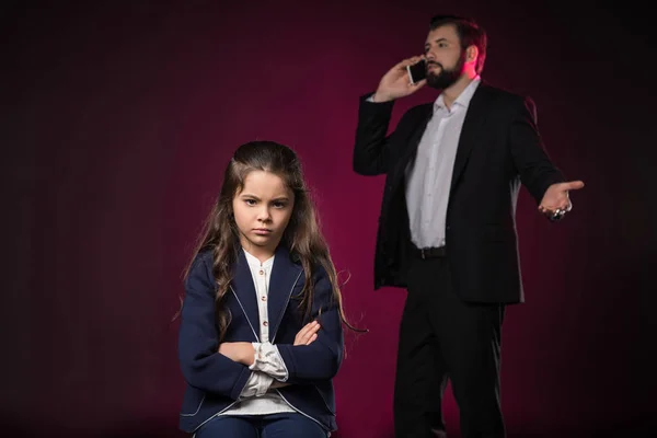 Hija enojada de pie con las manos cruzadas mientras el padre habla por teléfono inteligente en color burdeos - foto de stock