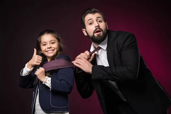 Daughter showing thumb up and pulling father tie on burgundy — Stock Photo