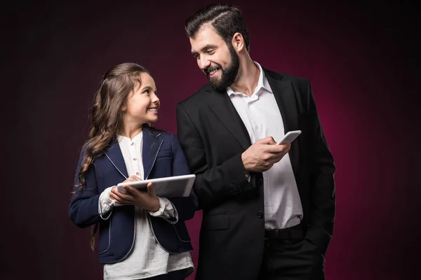 Smiling father and daughter with tablet and smartphone looking at each other on burgundy — Stock Photo