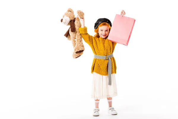 Feliz pelirroja niño sosteniendo bolsa de compras y juguete en las manos por encima de la cabeza aislado en blanco - foto de stock