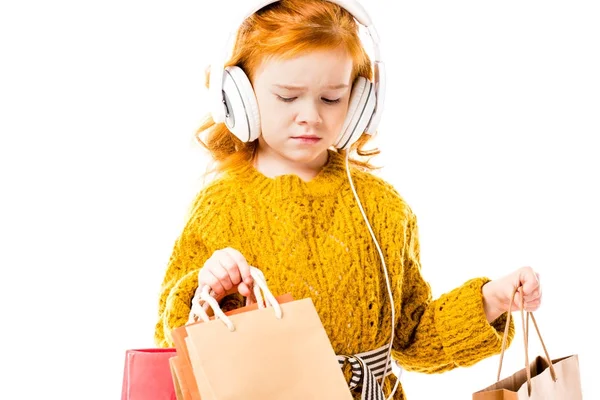 Niño molesto pelo rojo mirando las bolsas de la compra en las manos aisladas en blanco - foto de stock