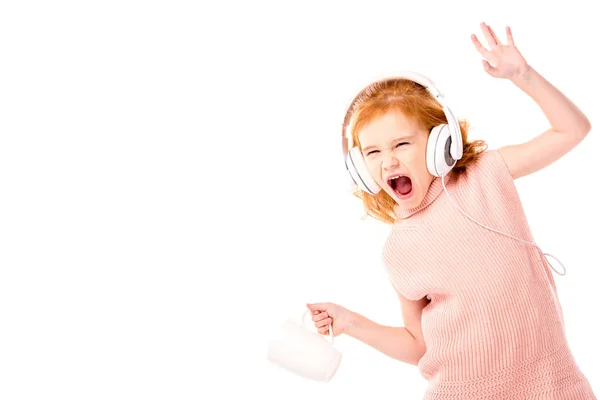 Cabrito de pelo rojo en auriculares gritando y bailando con taza aislada en blanco - foto de stock