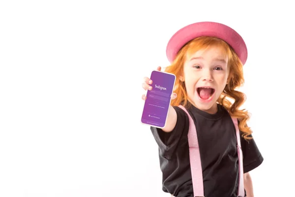 Elegante bambino in cappello rosa e bretelle mostrando smartphone con pagina instagram isolato su bianco — Foto stock