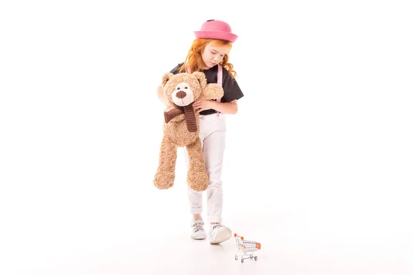 Aufgebrachtes Kind hält Teddybär in der Hand und schaut auf Einkaufswagen-Spielzeug auf weißem Grund — Stockfoto