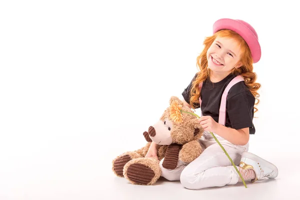 Lächeln rotes Haar Kind sitzt mit Teddybär und Blume auf weiß — Stockfoto