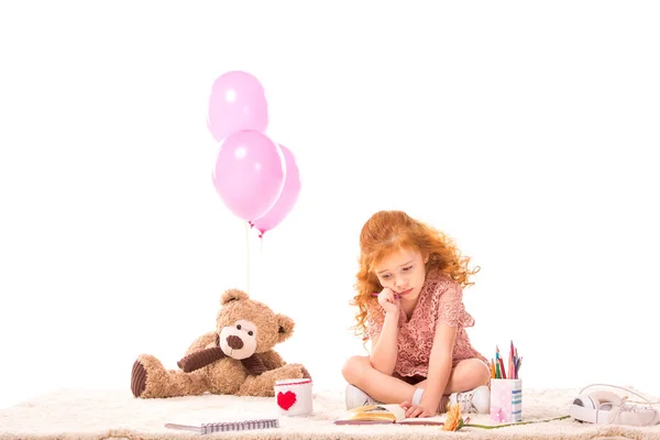 Сумне червоне волосся дитина сидить на килимі з олівцем і блокнотом ізольовано на білому — стокове фото