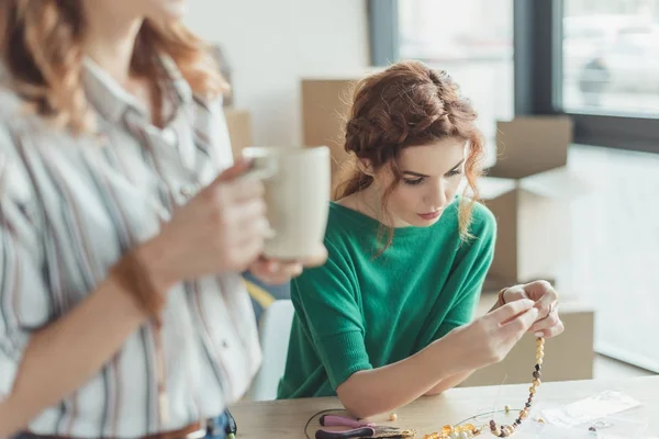 Konzentrierte junge Frau fertigt Accessoires in Werkstatt, während Kollegin Kaffee trinkt — Stockfoto