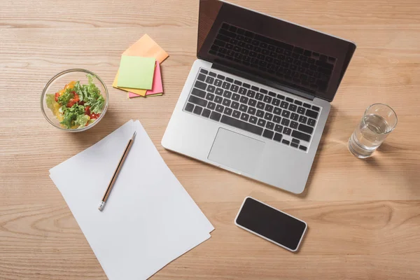 Vista superior del lugar de trabajo con computadora portátil, smartphone y deliciosa ensalada - foto de stock