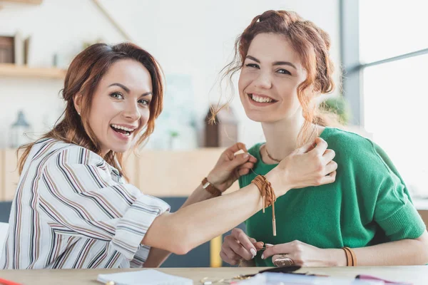 Sonrientes mujeres jóvenes probándose un collar hecho a mano en el taller - foto de stock