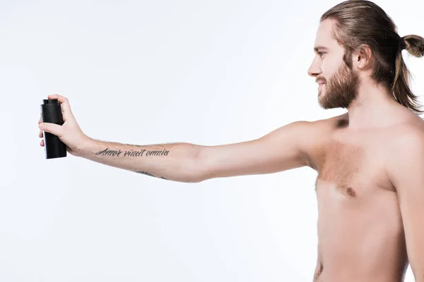 Immagine di giovane uomo dai capelli lunghi sorridente che tiene deodorante spray in mano tesa con tatuaggio, isolato su bianco — Foto stock