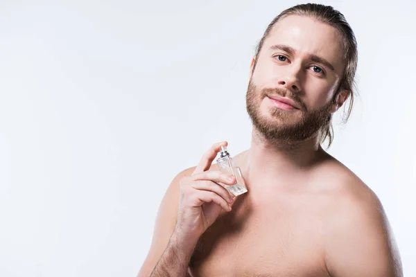 Barbuto giovane che guarda la macchina fotografica mentre tiene bottiglia di vetro con profumi contro il collo, isolato su bianco — Foto stock