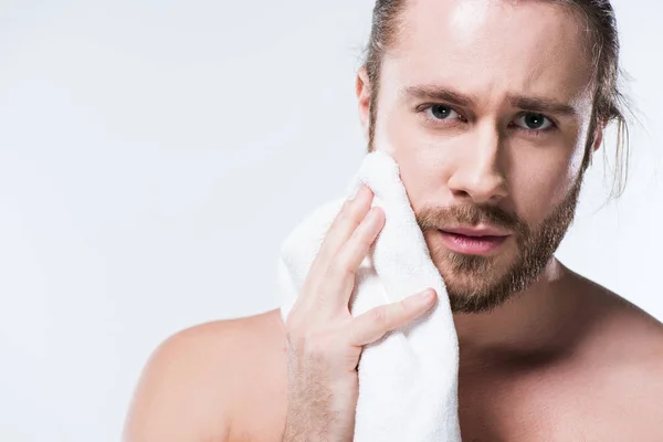 Молодий бородатий чоловік дивиться на камеру, тримаючи рушник для ванни в руці близько до щоки, ізольований на білому — стокове фото