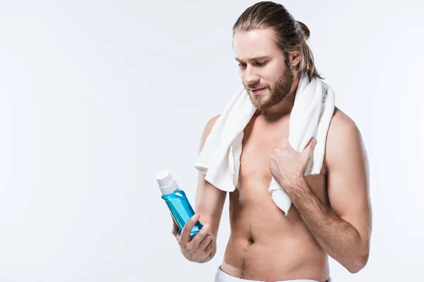 Smiling beardedman with towel around neck holding  tooth rinse in hand and looking on it, isolated on white — Stock Photo