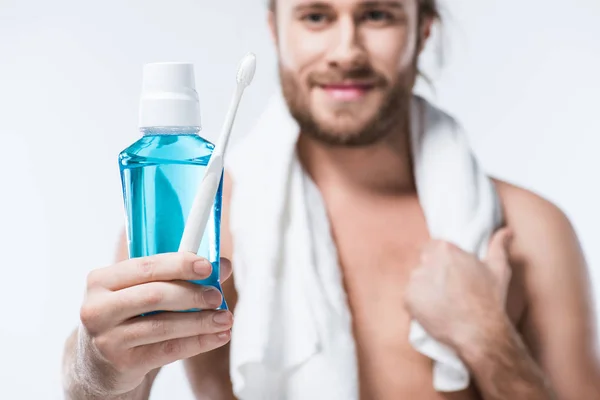 Smiling man with towel around his neck holding  tooth rinse and toothbrush in hand, isolated on white — Stock Photo