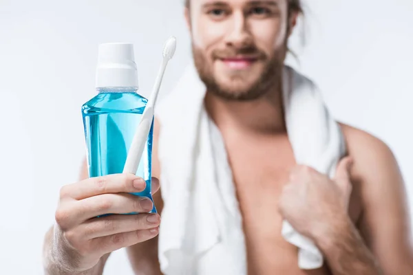 Hombre sonriente con toalla alrededor del cuello sosteniendo enjuague dental y cepillo de dientes en la mano, aislado en blanco - foto de stock