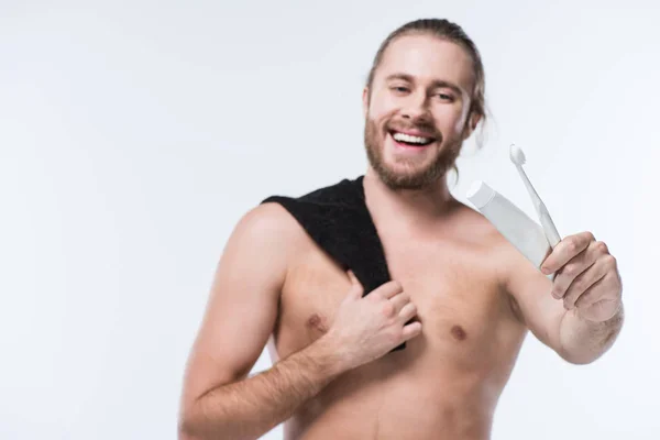 Jeune homme souriant avec une serviette noire sur l'épaule tenant du dentifrice et une brosse à dents à la main, isolé sur blanc — Photo de stock