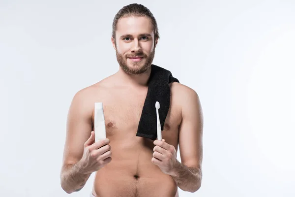 Cheerful young man with black towel on shoulder holding toothpaste in hand and toothbrush in another one,  isolated on white — Stock Photo