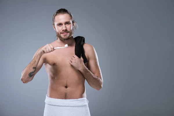 Man with black towel on shoulder holding toothbrush with toothpaste in hand against his face, isolated on gray — Stock Photo