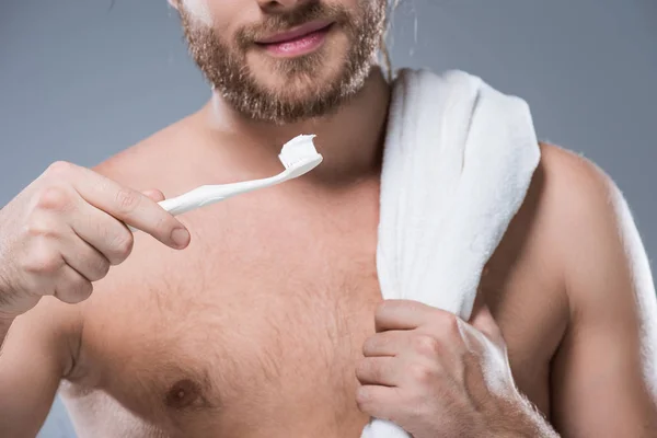 Imagen recortada de hombre barbudo sonriente con toalla en el hombro sosteniendo el cepillo de dientes con pasta de dientes en la mano, aislado en gris - foto de stock