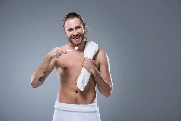 Joven sonriente con toalla de baño en el hombro sosteniendo el cepillo de dientes con pasta de dientes en la mano, aislado en gris - foto de stock