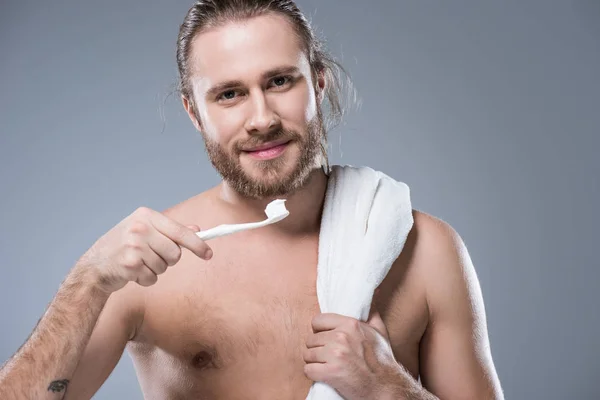 Smiling bearded man with towel on shoulder holding toothbrush with toothpaste in hand, isolated on gray — Stock Photo