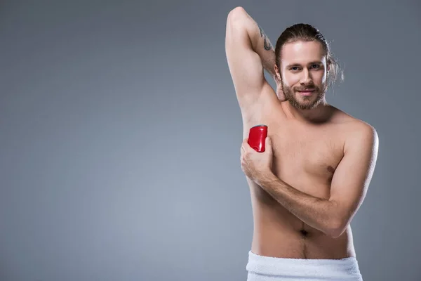 Giovane sorridente utilizzando bastone deodorante con mano dietro la testa, isolato su grigio — Foto stock