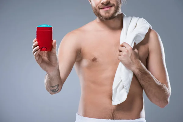 Smiling half naked man holding bath towel over shoulder in one hand and deodorant stick in another, isolated on gray — Stock Photo